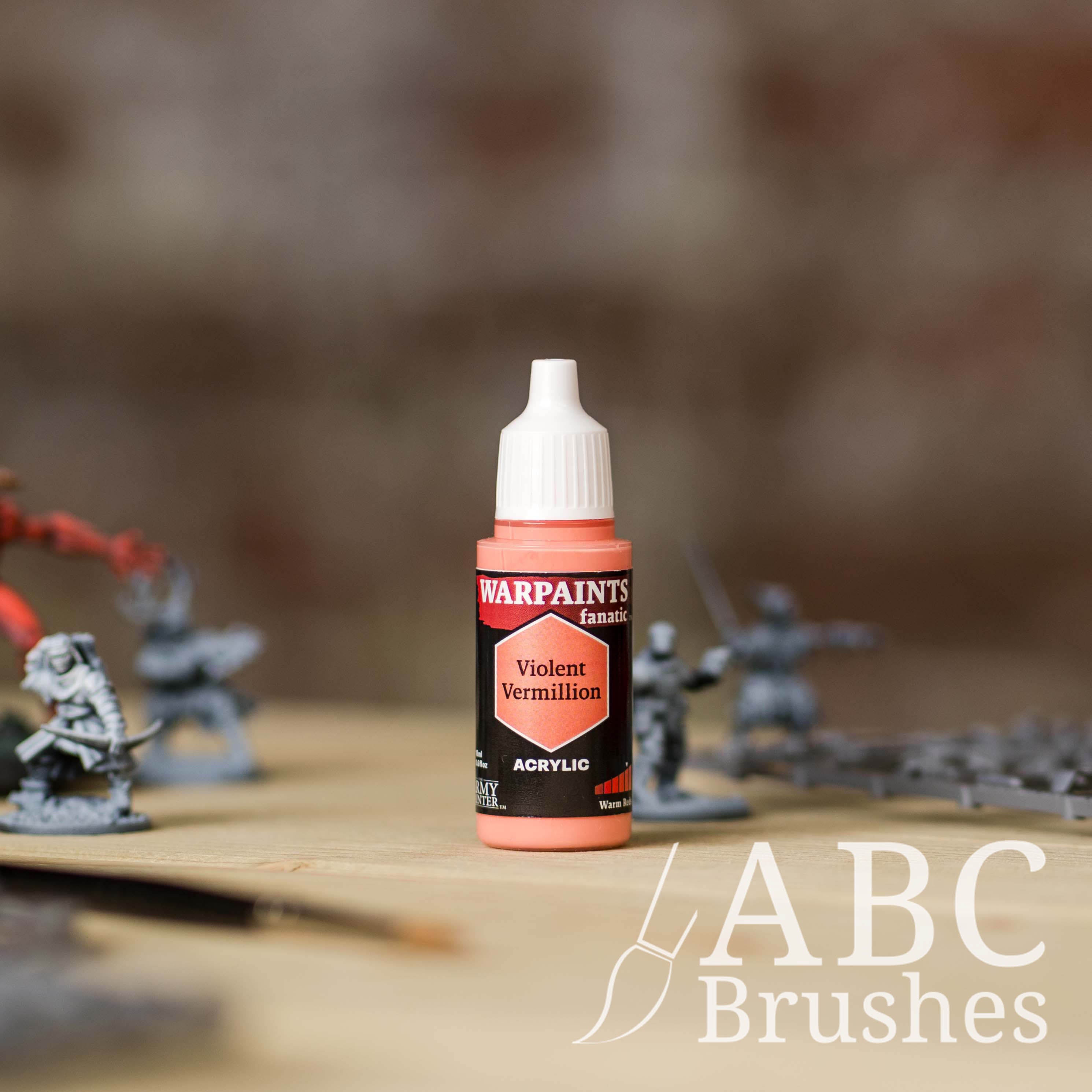 Bottle of the Army Painter Fanatics in the colour Violent Vermillion with various wargaming models and an ABC paintbrush in the background, on a wooden surface.