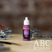Bottle of the Army Painter Fanatics in the colour Warlock Magenta with various wargaming models and an ABC paintbrush in the background, on a wooden surface.
