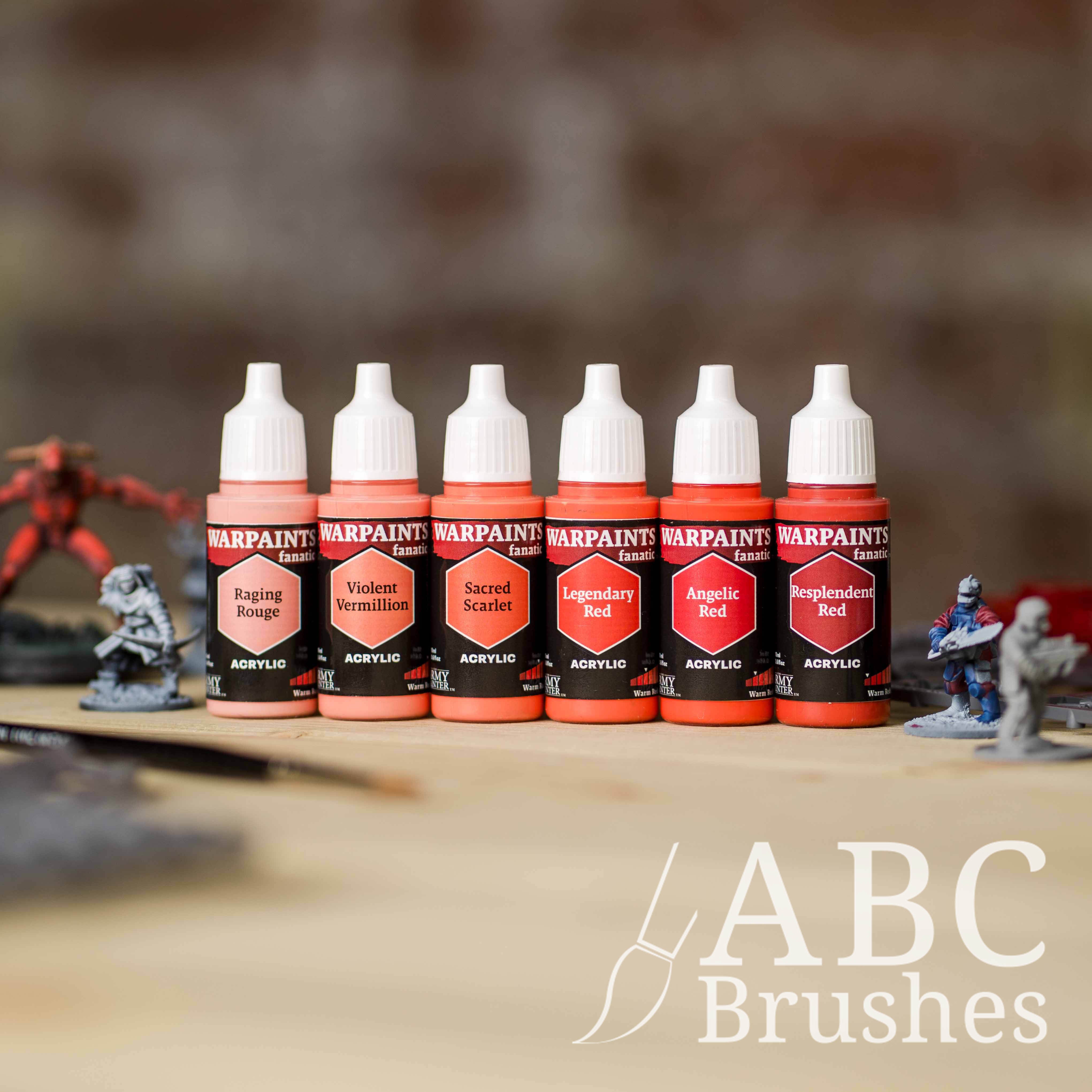 Army Painter Fanatics Warm Reds Triad on a wooden surface. There are wargaming models and an ABC paint brush next to them.