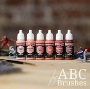 Army Painter Fanatics Warm Skin Tone Triad on a wooden surface. There are wargaming models and an ABC paint brush next to them.