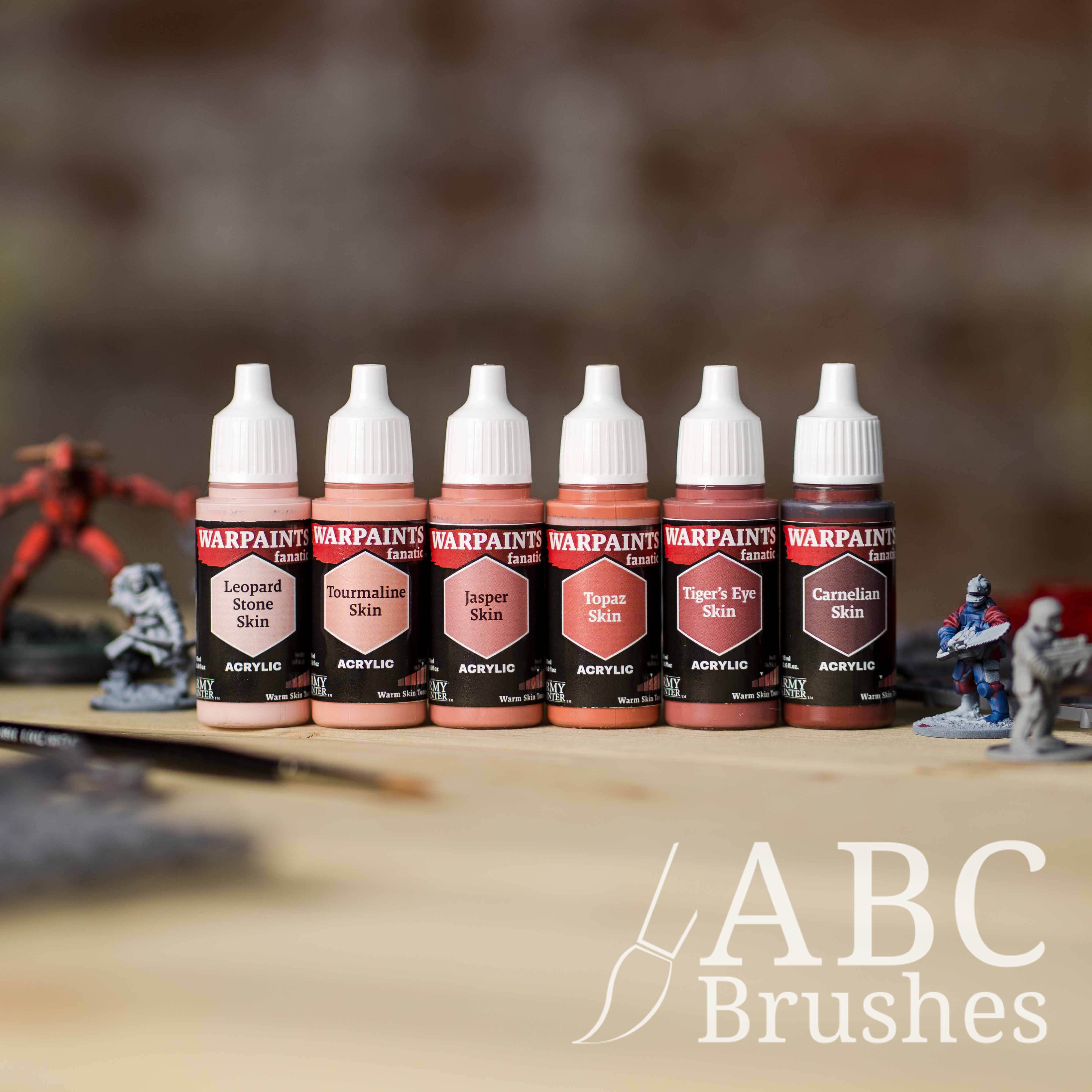Army Painter Fanatics Warm Skin Tone Triad on a wooden surface. There are wargaming models and an ABC paint brush next to them.