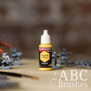 Bottle of the Army Painter Fanatics in the colour Warped Yellow with various wargaming models and an ABC paintbrush in the background, on a wooden surface.