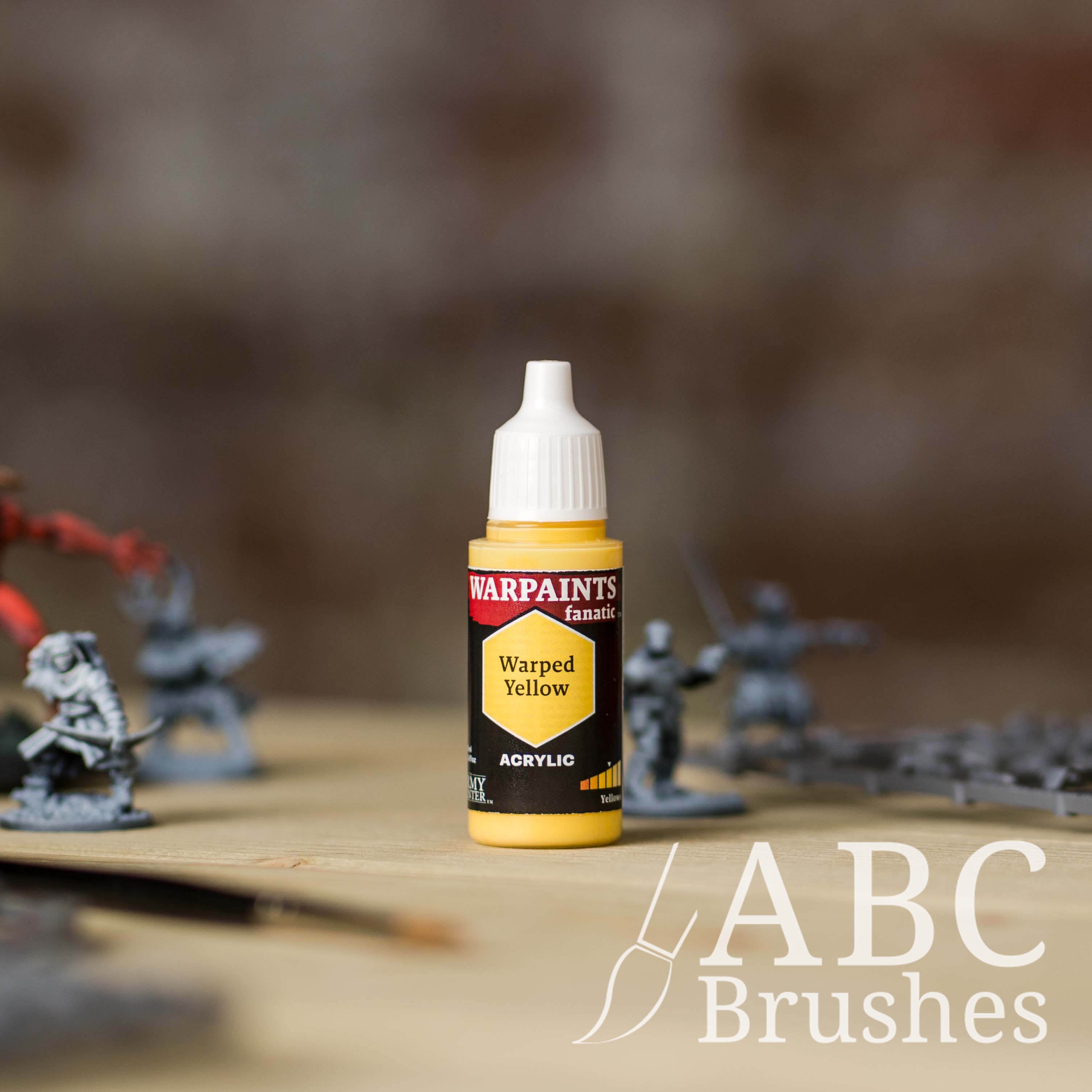 Bottle of the Army Painter Fanatics in the colour Warped Yellow with various wargaming models and an ABC paintbrush in the background, on a wooden surface.
