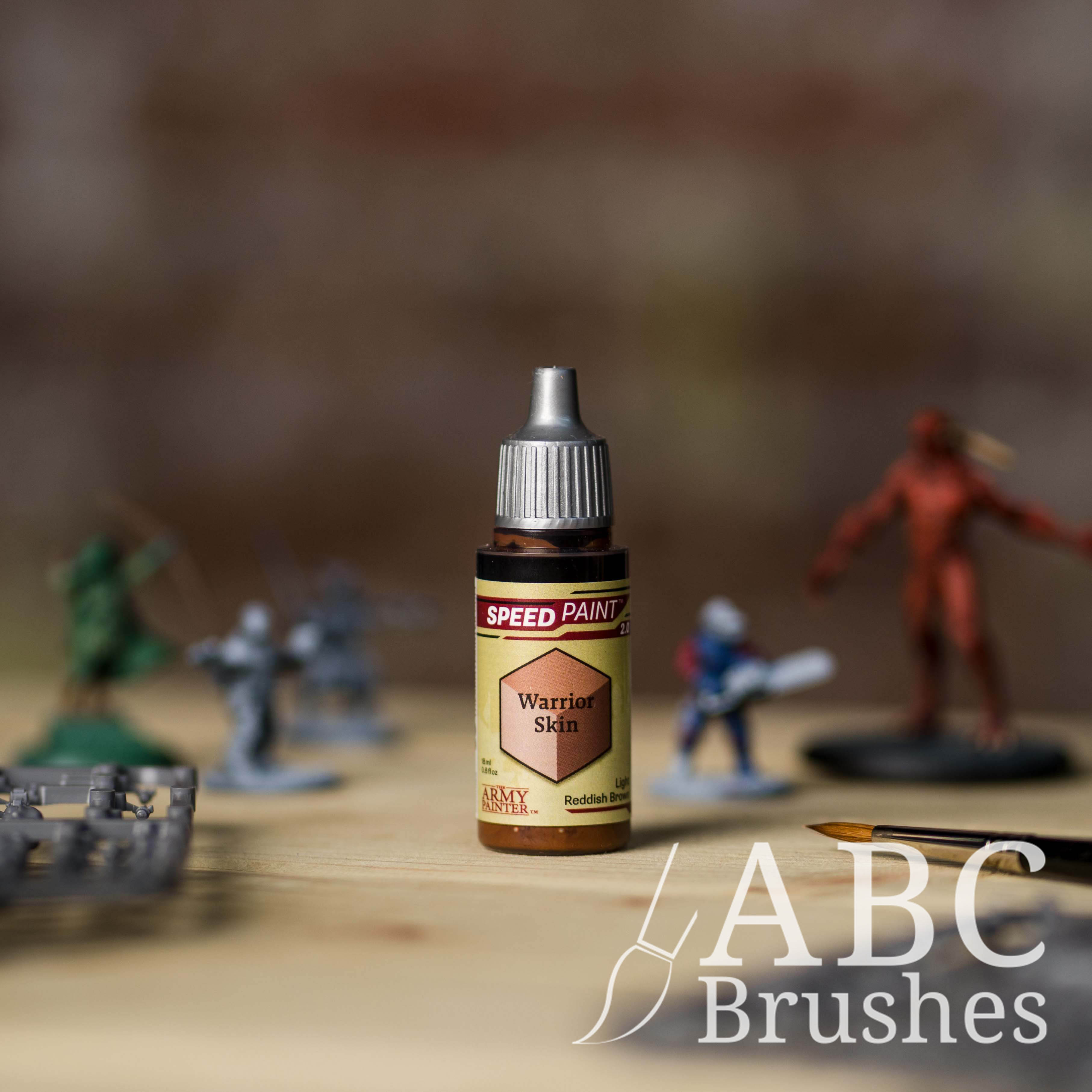 Bottle of the Army Painters Speedpaint in the colour Warrior Skin, with wargaming models and an ABC paintbrush in the background, on a wooden surface.