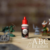 Bottle of the Army Painter Fanatics Wash Medium with various wargaming models and an ABC paintbrush in the background, on a wooden surface.
