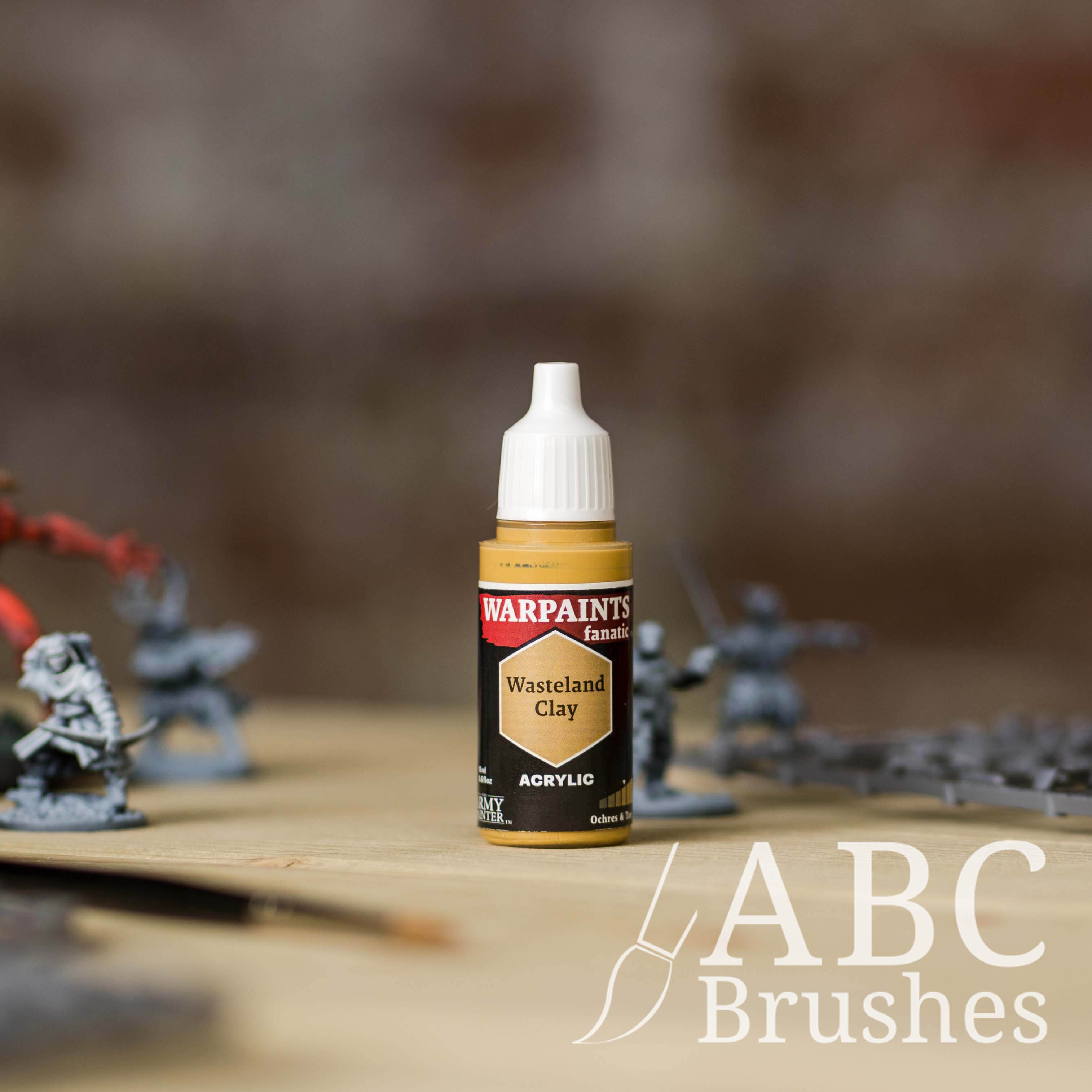 Bottle of the Army Painter Fanatics in the colour Wasteland Clay with various wargaming models and an ABC paintbrush in the background, on a wooden surface.