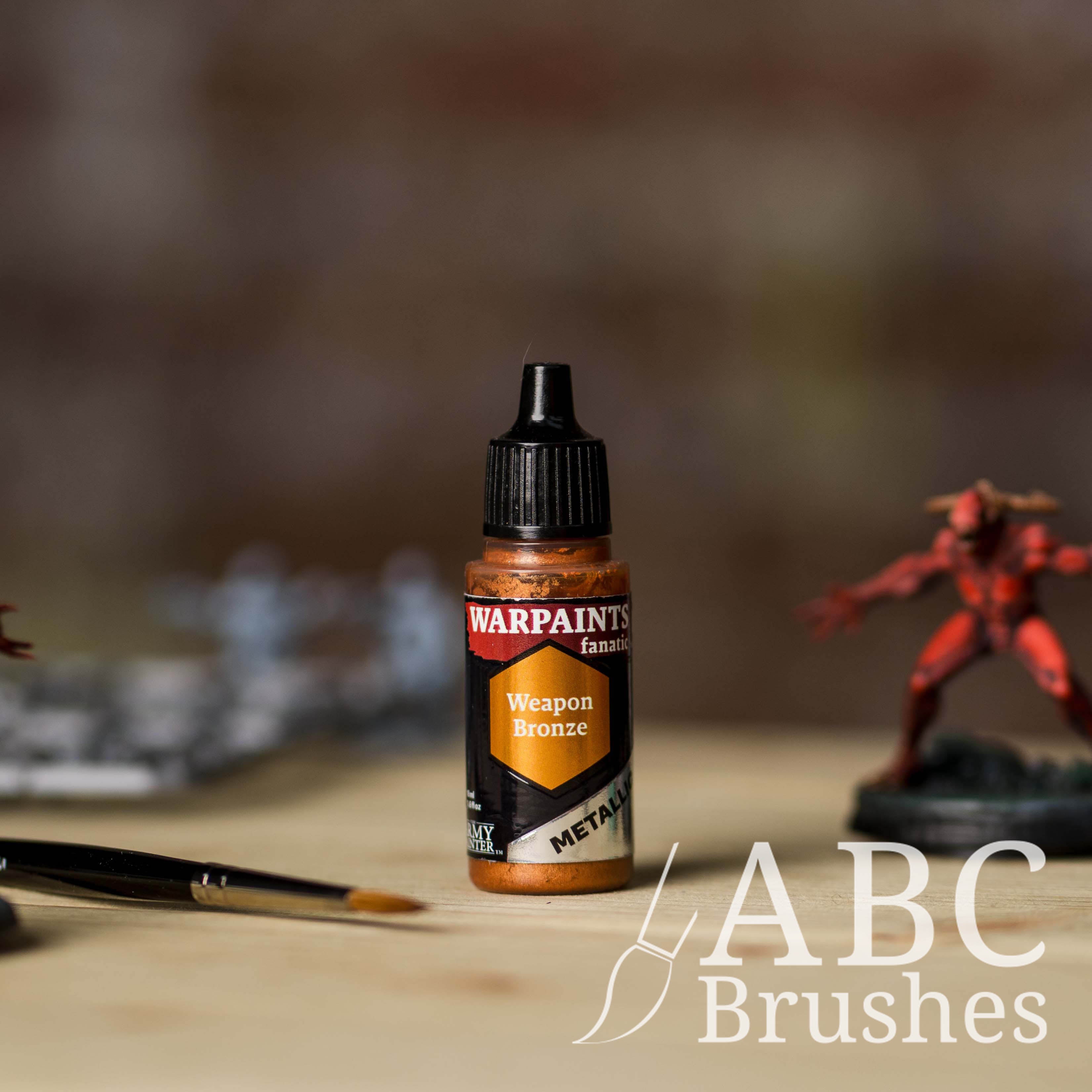 Bottle of the Army Painter Fanatics Metallic paint in the colour Weapon Bronze with various wargaming models and an ABC paintbrush in the background, on a wooden surface.