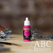 Bottle of the Army Painter Fanatics in the colour Wicked Pink with various wargaming models and an ABC paintbrush in the background, on a wooden surface.