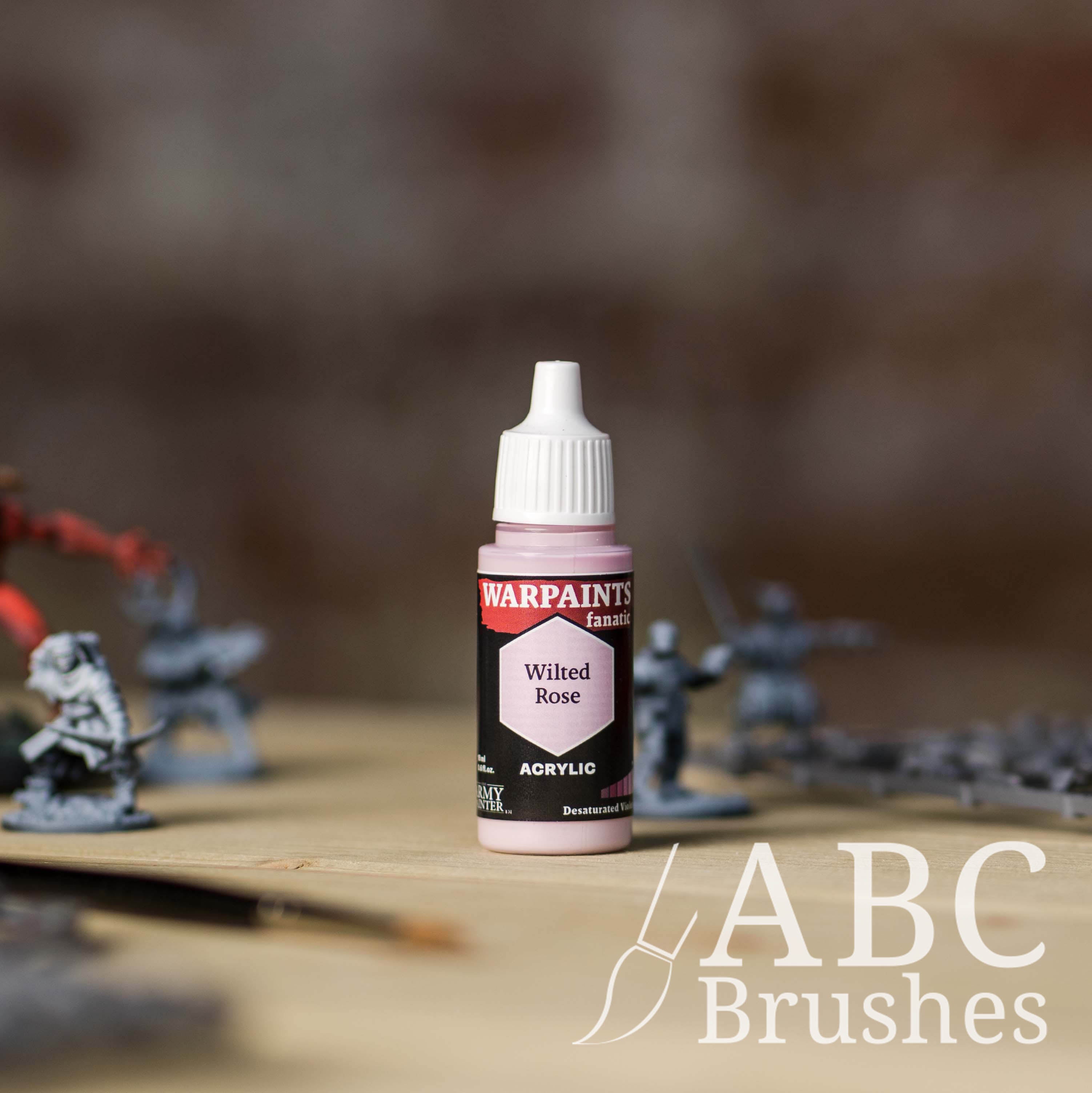 Bottle of the Army Painter Fanatics in the colour Wilted Rose with various wargaming models and an ABC paintbrush in the background, on a wooden surface.