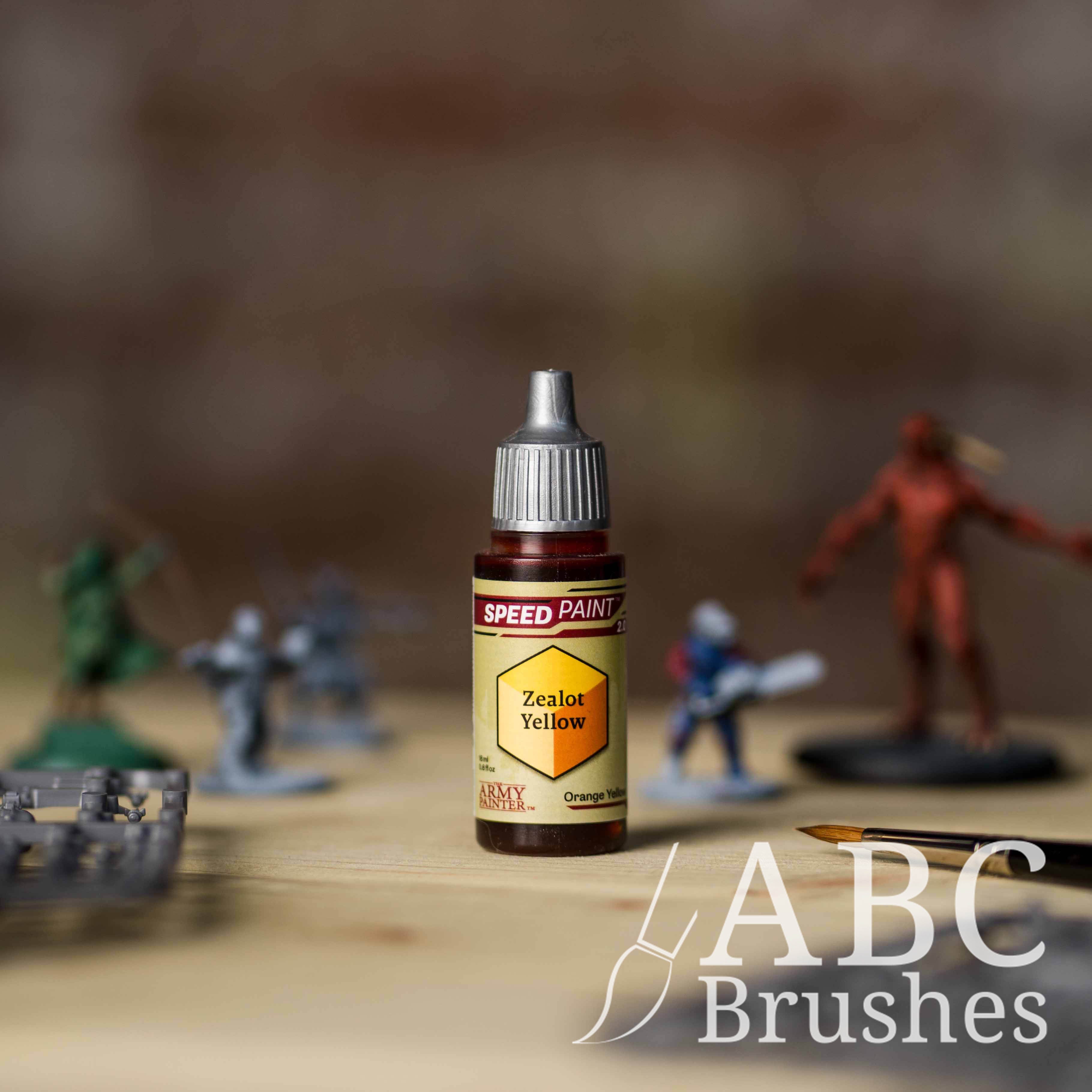 Bottle of the Army Painters Speedpaint in the colour Zealot Yellow, with wargaming models and an ABC paintbrush in the background, on a wooden surface.