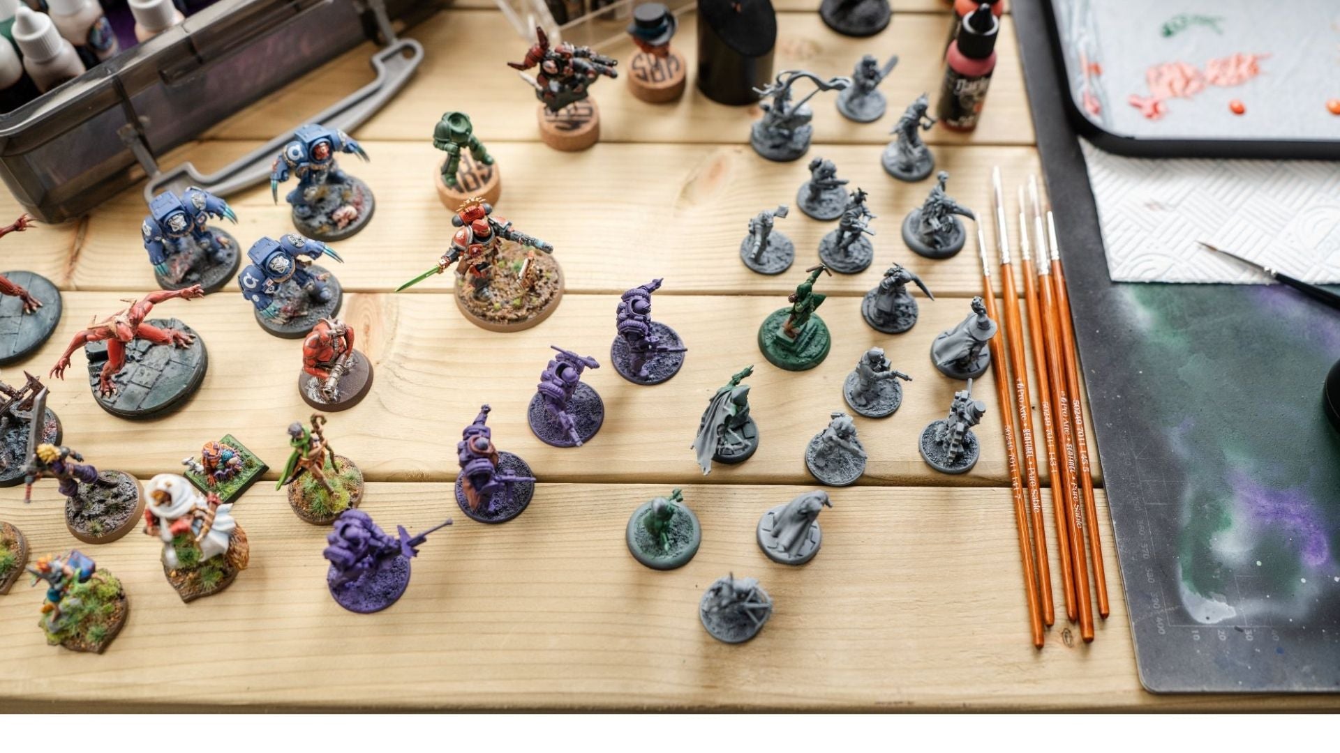 Collection of mini figures on a wooden table with various paintbrushes, paints and other supplies. 