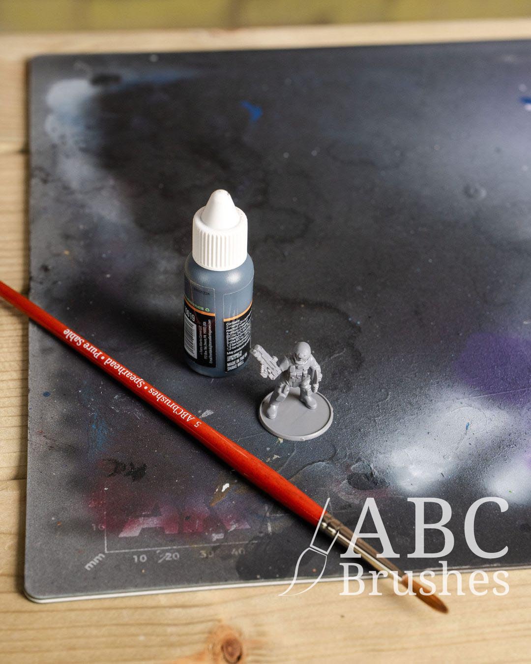 ABC Sentinel round paintbrush, bottle of acrylic paint, and a miniature figure on a paint covered cutting mat surface with a wooden background.