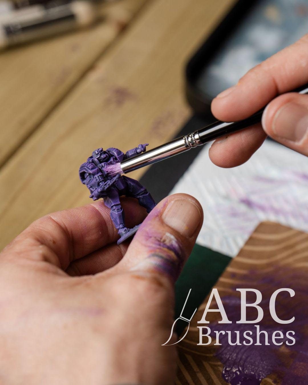 Painting a purple wargaming mini with a Pro Arte dry brush. In the background you can see a wet palette and selection of Pro Arte dry brushes. 