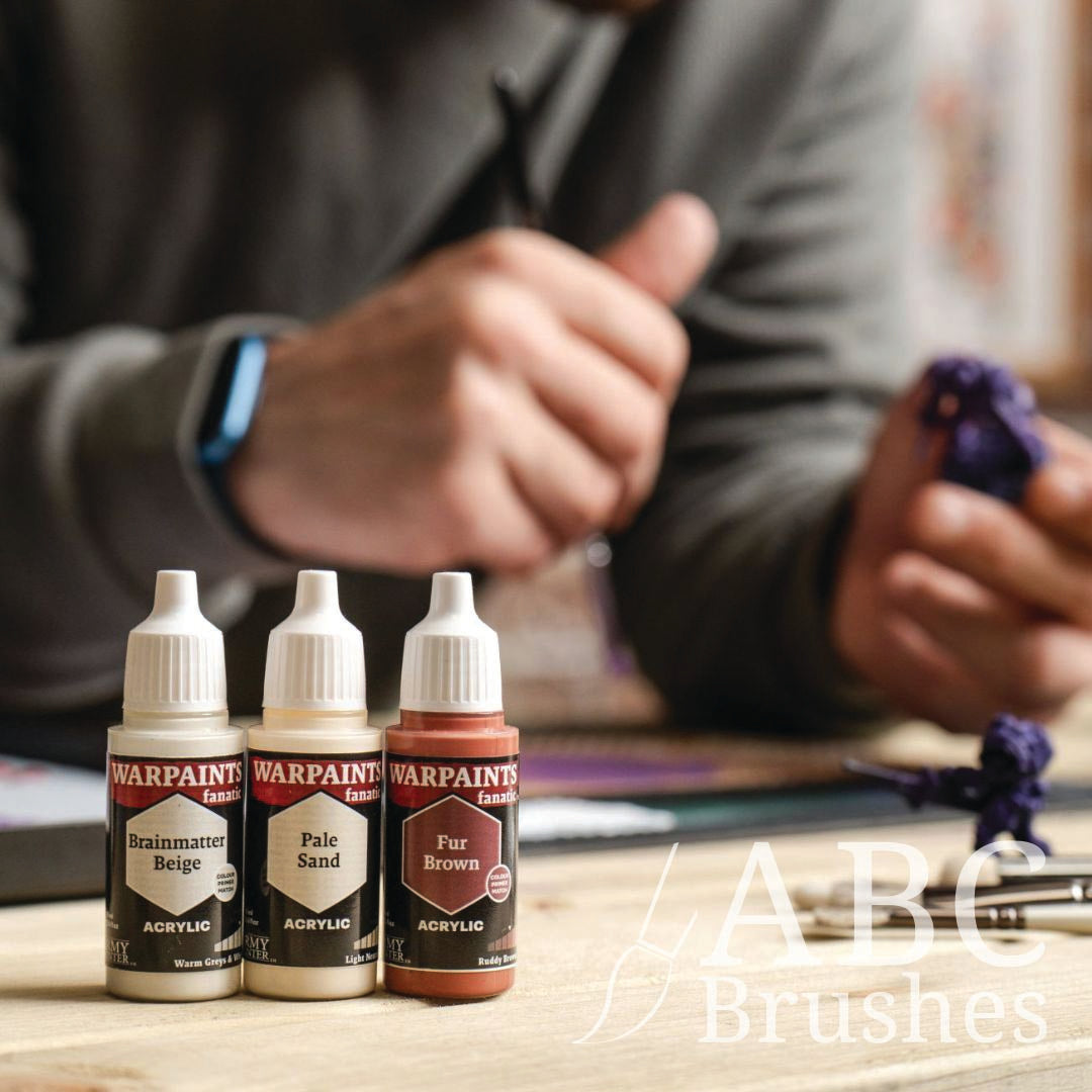 Three bottles of War Paints Fanatics model paint on a table with a person working on a model in the background.