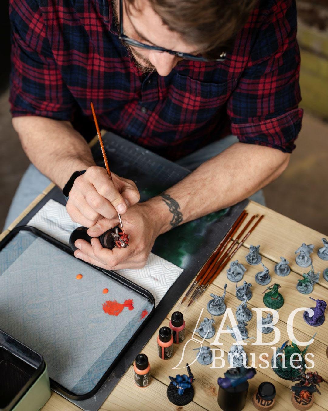  BlackHeartMinis painting a mini with an ABC Kolinsky Sable brush. There is a wet palette and various paint and models on the table below him. 