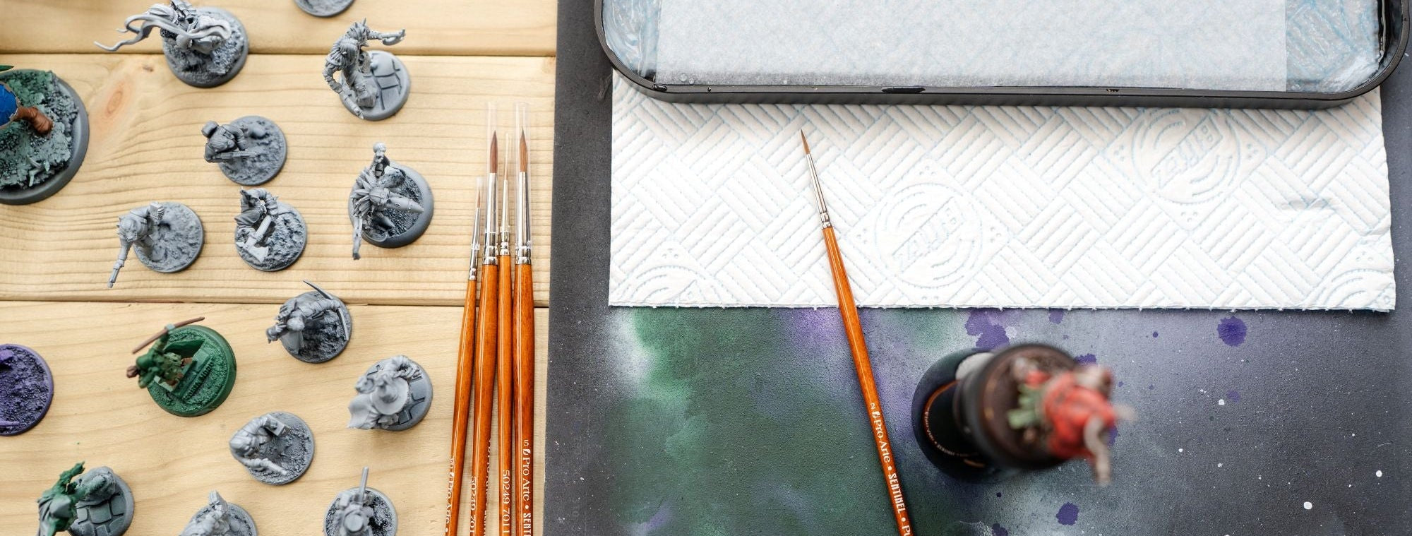 Miniatures, ABC Sentinel paintbrushes, wet palette and a paint covered cutting mat set up on a wooden surface. 