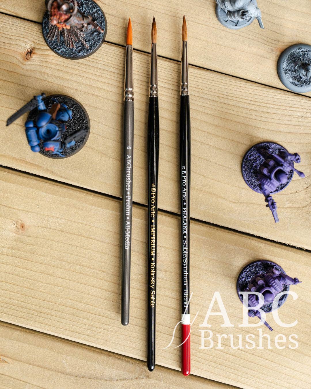 Three Pro Ate and ABC paint brushes, Prolon, Imperium and Phalanx arranged next to each other on a wooden surface. There are various wargaming minis surrounding them. 